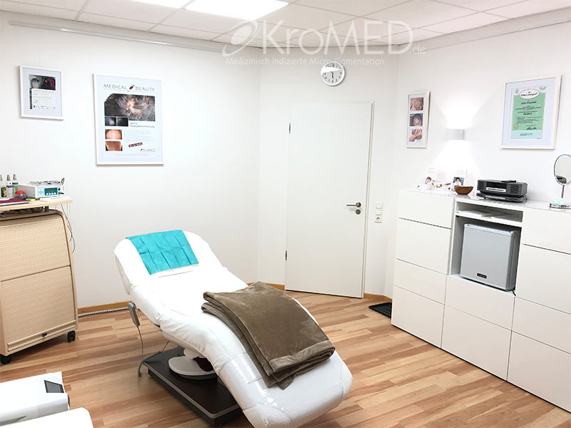 KroMED | Bild Behandlungsraum- Praxis für micro Haarpigmentierung und Permanent Make-up- Hamburg. Das Bild zeigt einen weiteren Bereich eines Kosmetikstudios, der ebenfalls hell und modern gestaltet ist. Im Mittelpunkt steht eine Behandlungsliege mit einem blauen Kopfkissen und einer braunen Decke. Links daneben befindet sich ein rollbarer Schrank mit diversen Kosmetikprodukten und Geräten darauf. Die Wände sind mit verschiedenen gerahmten Bildern und Zertifikaten dekoriert, die die Professionalität des Studios unterstreichen. An der rechten Wand steht ein weißes Sideboard, das einen kleinen Kühlschrank und zusätzliche Aufbewahrungsfächer integriert. Eine Wandleuchte sorgt für angenehme Beleuchtung. Ein weißer Türrahmen und ein Holzboden vervollständigen das harmonische Ambiente des Raums.