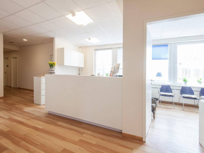 KroMED | Empfang Praxis für aesthetische Pigmentierung und Permanent Make-up- Hamburg. Der Eingang zur Praxis KroMED wirkt hell und freundlich. Der Raum ist in warmen, neutralen Tönen gestaltet, die durch das helle Holzlaminat des Bodens betont werden. Ein Empfangstresen in sanftem Cremeweiß begrüßt die Patienten direkt beim Betreten und verleiht dem Raum eine einladende Atmosphäre. Über dem Tresen hängen kleine, moderne Lampen, die eine dezente Beleuchtung bieten. An der Wand rechts hängt ein großformatiges Bild mit roten Rosen, was einen freundlichen und beruhigenden Farbakzent setzt. Die Wände sind in einem warmen Beige gestrichen, was zur ruhigen Ausstrahlung beiträgt. Es gibt auch Spiegel, die den Raum optisch vergrößern. Hinter dem Empfangstresen befinden sich Regale und Schränke, die ordentlich und unaufdringlich wirken. Ein kleiner Tisch mit Zeitschriften und Dekorationen bietet den Patienten einen Wartebereich in angenehmer Umgebung.