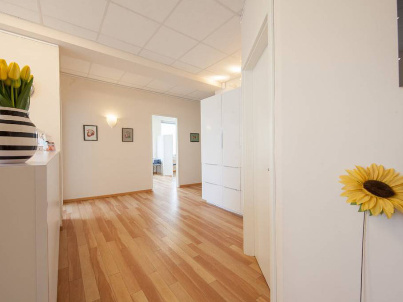 KroMED | Bild Eingangsbereich - Praxis für aesthetische Pigmentierung und Permanent Make-up- Hamburg. Der Empfangsbereich der KroMED-Praxis ist in hellen Farben und mit einem warmen Holzlaminatboden gestaltet, was eine freundliche und beruhigende Atmosphäre schafft. Der Empfangstresen befindet sich links im Bild und wirkt modern und einladend. Links neben dem Empfang führt eine Glastür zu einem weiteren Raum. Der Blick geht durch eine offene Tür geradeaus in das Behandlungszimmer, wo spezialisierte Behandlungen wie Permanent Make-up (PMU), Diamant Blading, Brustwarzenpigmentierung und micro Haarpigmentierung angeboten werden. Im Behandlungsraum selbst ist eine professionelle und entspannte Umgebung zu erkennen. Der Raum wirkt gut organisiert und einladend, mit geschmackvoller Dekoration und hochwertigen Behandlungsgeräten. Die Wände sind in einem warmen Ton gehalten, was die ruhige Atmosphäre der Praxis unterstreicht.